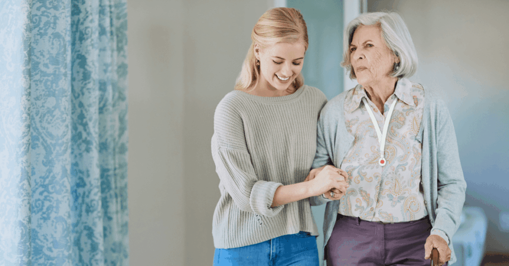 Eine junge Frau unterstützt eine ältere Dame beim Gehen – beide lächeln. Die Szene zeigt den liebevollen Alltag von pflegende Angehörige. Das Bild steht im Kontext der IAHA – International Association for Healthy Aging, die pflegende Angehörige mit Beratung und Schulung gezielt unterstützt.